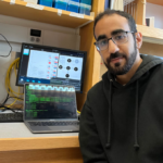 A man with short dark hair, a beard, and glasses sits at a lab workstation. He wears a black hoodie and looks toward the camera with a slight smile. Behind him are two computer screens—one showing a terminal with lines of code and the other displaying a flowchart with scientific diagrams. Shelves above the desk hold plastic lab containers labeled “200 uL Filter” and “1000 uL Filter.”