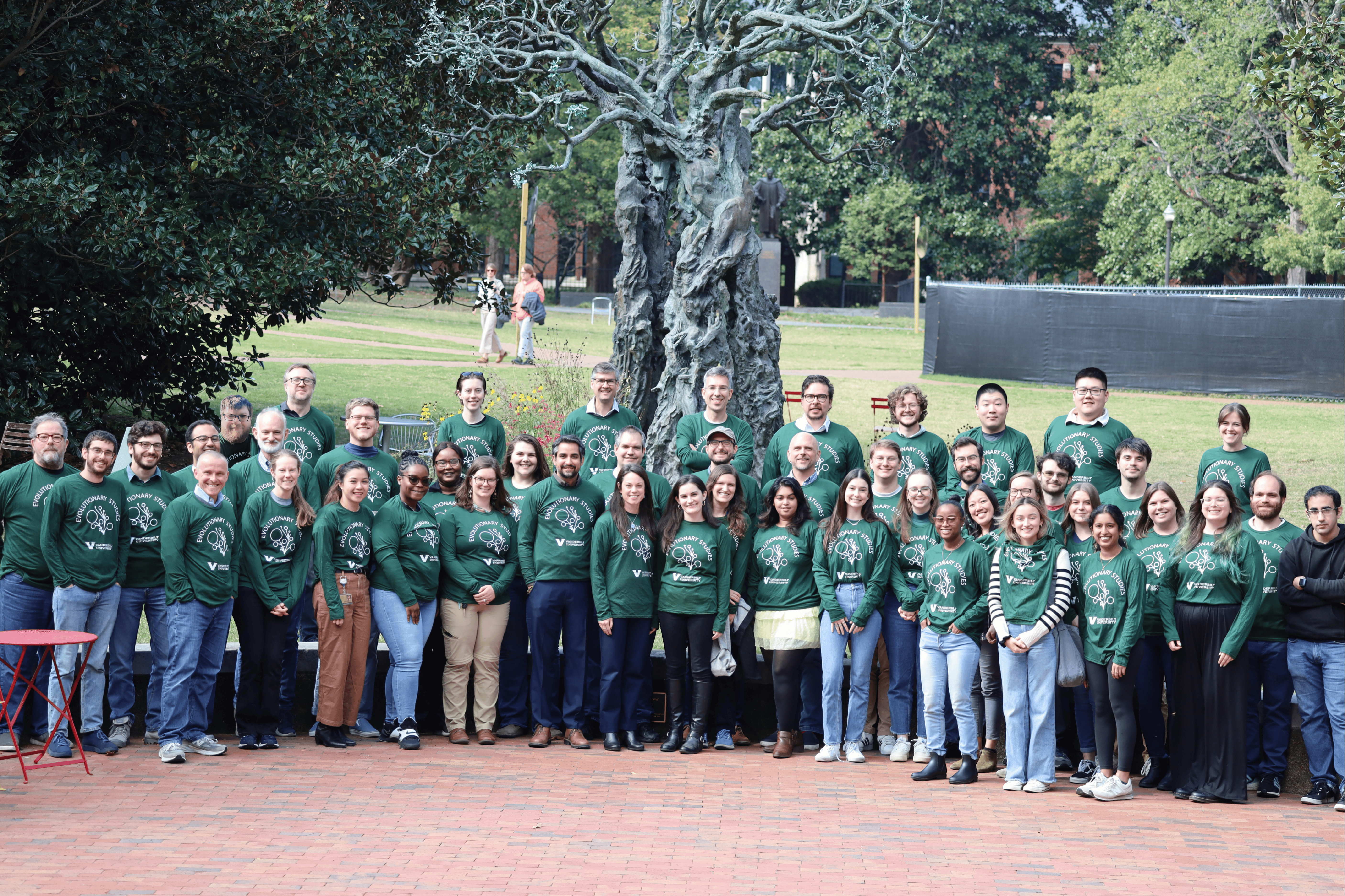 A large group of people all wearing dark green, long-sleeved shirt smiling at a camera. They stand in front of a sculpture of a tree. The ground is red brick in front and grass in the background.